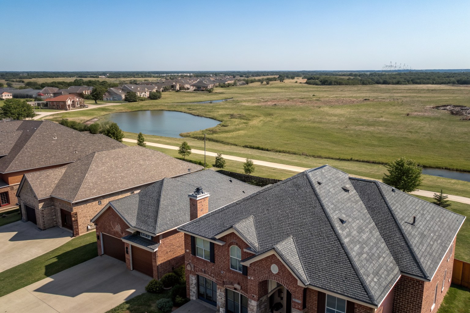 Aerial view of brick homes with gray and brown shingle roofs beside a pond on open prairie
