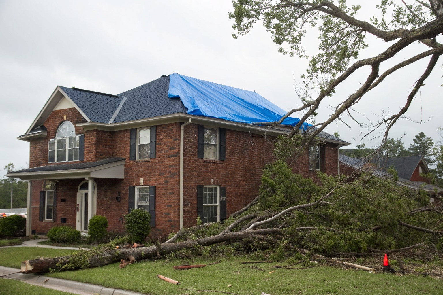 A two-story brick home with a blue tarp over part of its roof and broken limbs on the lawn